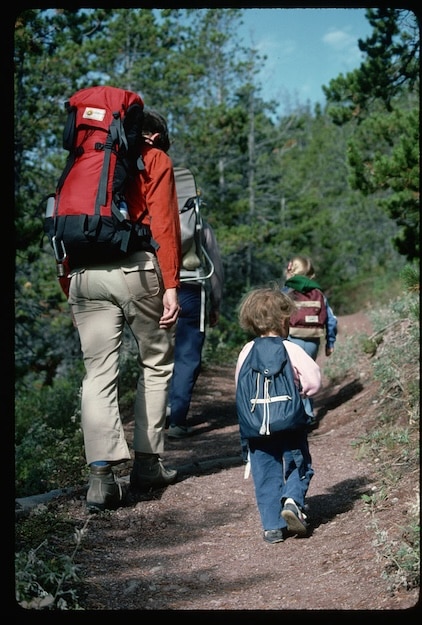 An adult and two kids backpacking with backpacks hike along a dirt trail through a forested area, surrounded by trees and lush greenery.