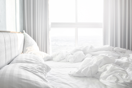 A white bed with freshly made white sheets and pillows in front of a window.