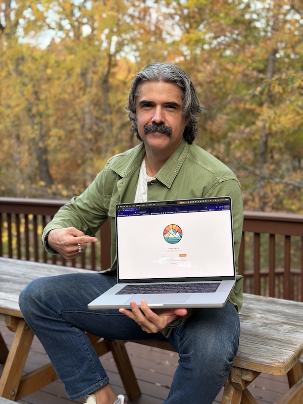 A man sits on a bench outdoors, holding a laptop with the Art of Manliness logo displayed on the screen and pointing at it. Autumn trees are visible in the background, suggesting support for their free newsletter.