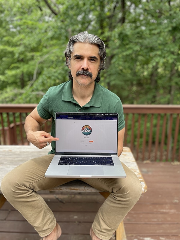 A man sitting on a wooden bench outdoors points to the screen of an open laptop displaying the Dying Breed website with its circular logo.