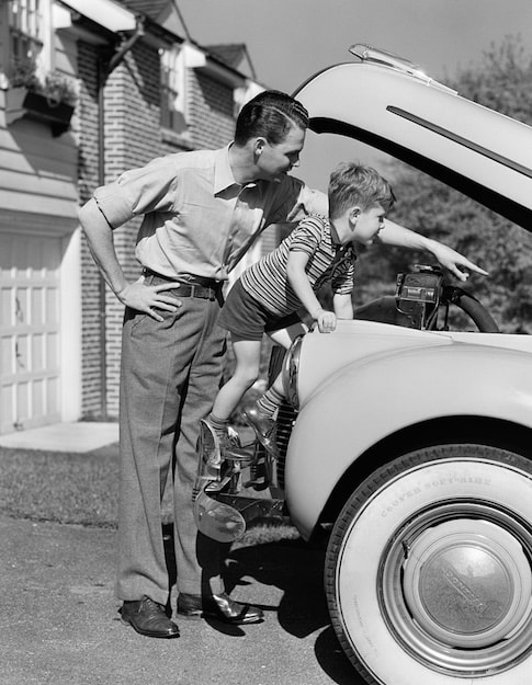 A man in classic men’s style and a young boy look under the hood of a car parked in a driveway. The man stands while the boy stands on the fender, both focused on the engine.