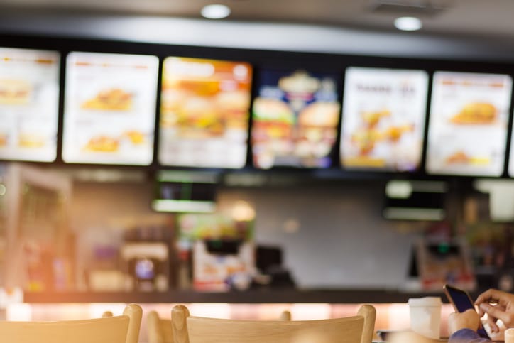 Blurry image of a fast food restaurant counter with illuminated menu boards offering meals less than 600 calories and high in protein, and a person using a phone in the foreground.