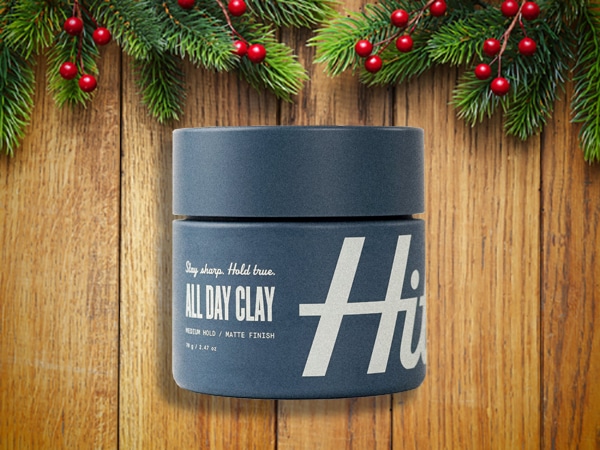 A jar of hair styling clay labeled "All Day Clay" sits on a wooden surface, surrounded by festive green branches and red berries, making it the perfect stocking stuffer for men.