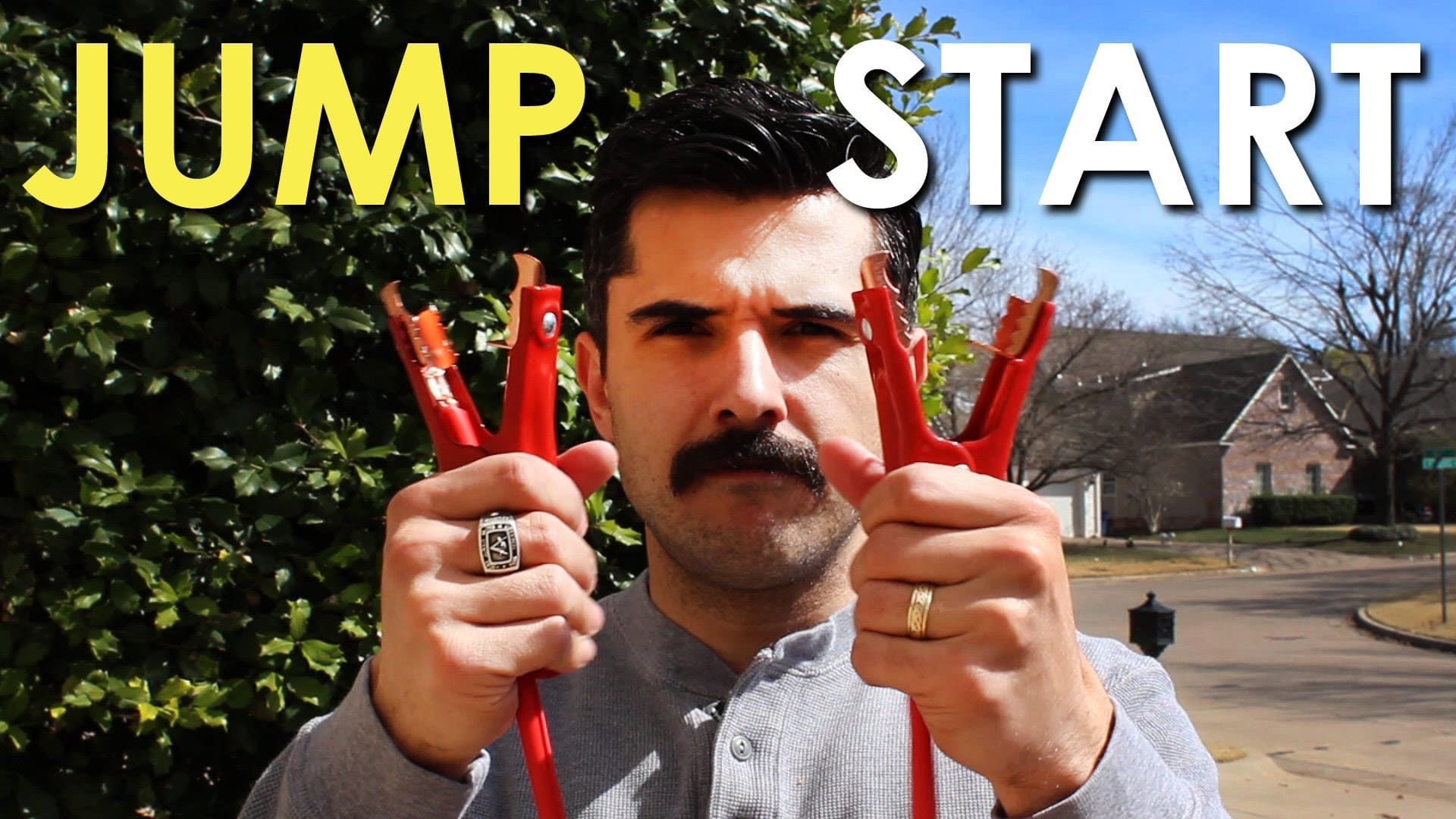 A man with a mustache is holding a pair of jumper cables next to a car he is about to jump start.