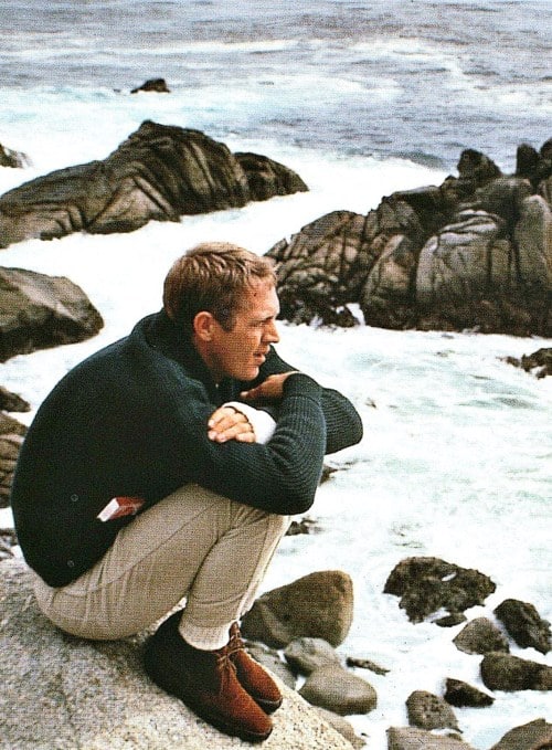 Steve mcqueen sitting on ocean rocks.