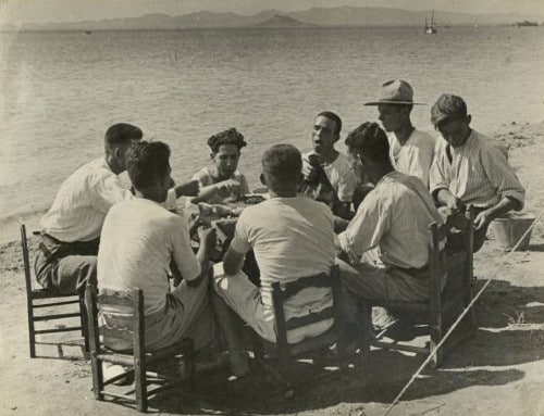Vintage men eating around dinner table on beach ocean.