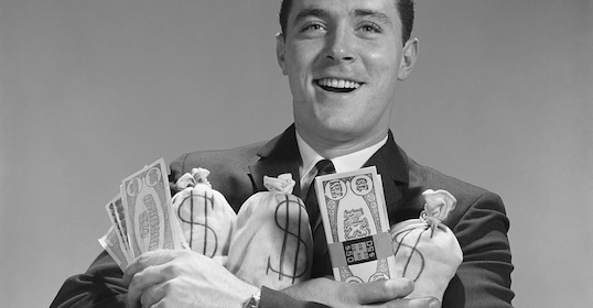 A smiling man in his 30s in a suit holds several bags marked with dollar signs and large paper bills, celebrating his personal finance goals.