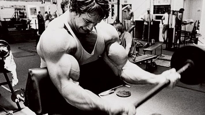A person in a gym intensely performs a bicep curl with a barbell on a preacher bench. Other gym-goers and equipment are visible in the background.