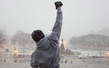 A man in a hoodie raises his fist in the snow, as if ready to engage in a boxing match.