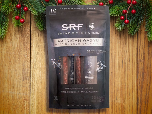A package of SRF Snake River Farms American Wagyu beef smoked sausages sits on a wooden surface with evergreen branches decorating the edges, making it an ideal choice for stocking stuffers for men this holiday season.