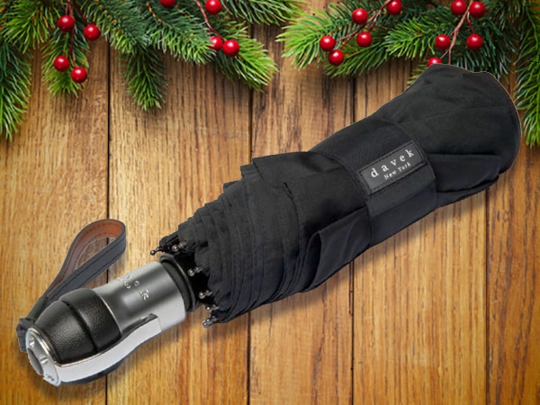 A closed black Davek umbrella with a wrist strap rests on a wooden surface, surrounded by evergreen branches and red berries—an ideal pick for stocking stuffers for men.