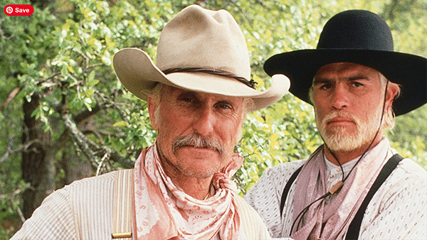 Two men in Western attire, resembling Gus and Woodrow, stand outdoors in front of leafy green trees, both wearing wide-brimmed hats and bandanas.